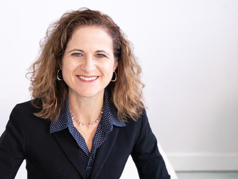 Woman with brown hair wearing black jacket in front of white wall